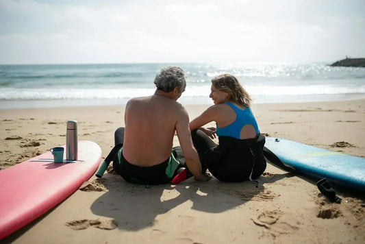A man and woman sit on a sandy beach, enjoying a sunny day with surfboards and a thermos