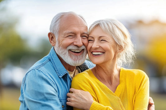 A senior couple in a loving embrace in warm sunlight in front of a blurry nature background, both smiling.