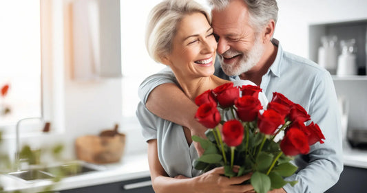 A woman with gray hair holding a bouquet of red roses and smiling as a man with gray hair smiles at her while embracing her from behind.